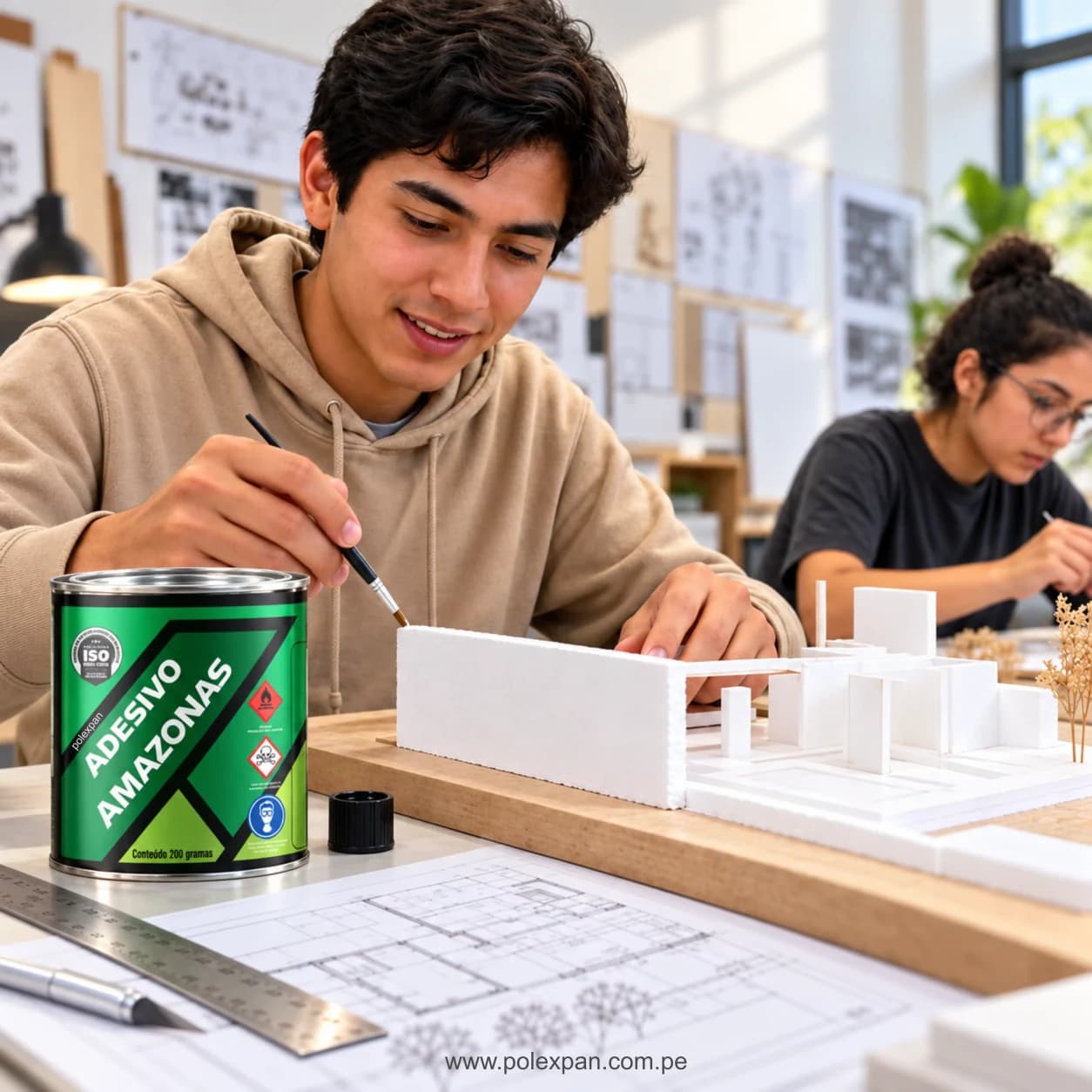 Estudiante pegando piezas de tecnopor en una maqueta de arquitectura, con planos sobre la mesa y otra estudiante trabajando al fondo; en primer plano, la lata de adhesivo Amazonas rotulada «Conteúdo 200 gramas».